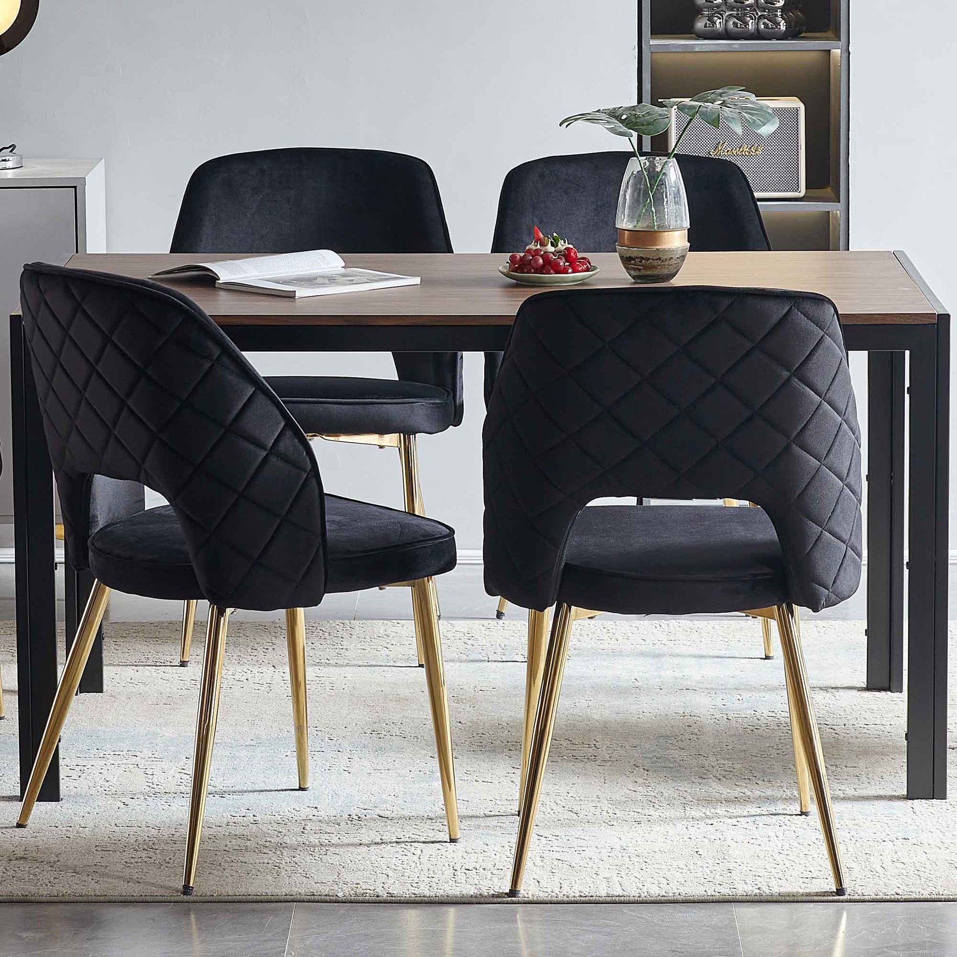 A modern dining area features a wooden table and four Velvet Dining Chairs With Metal Legs And Hollow Back, plus a small plant in a vase, an open book, and a plate of cherries on the table.