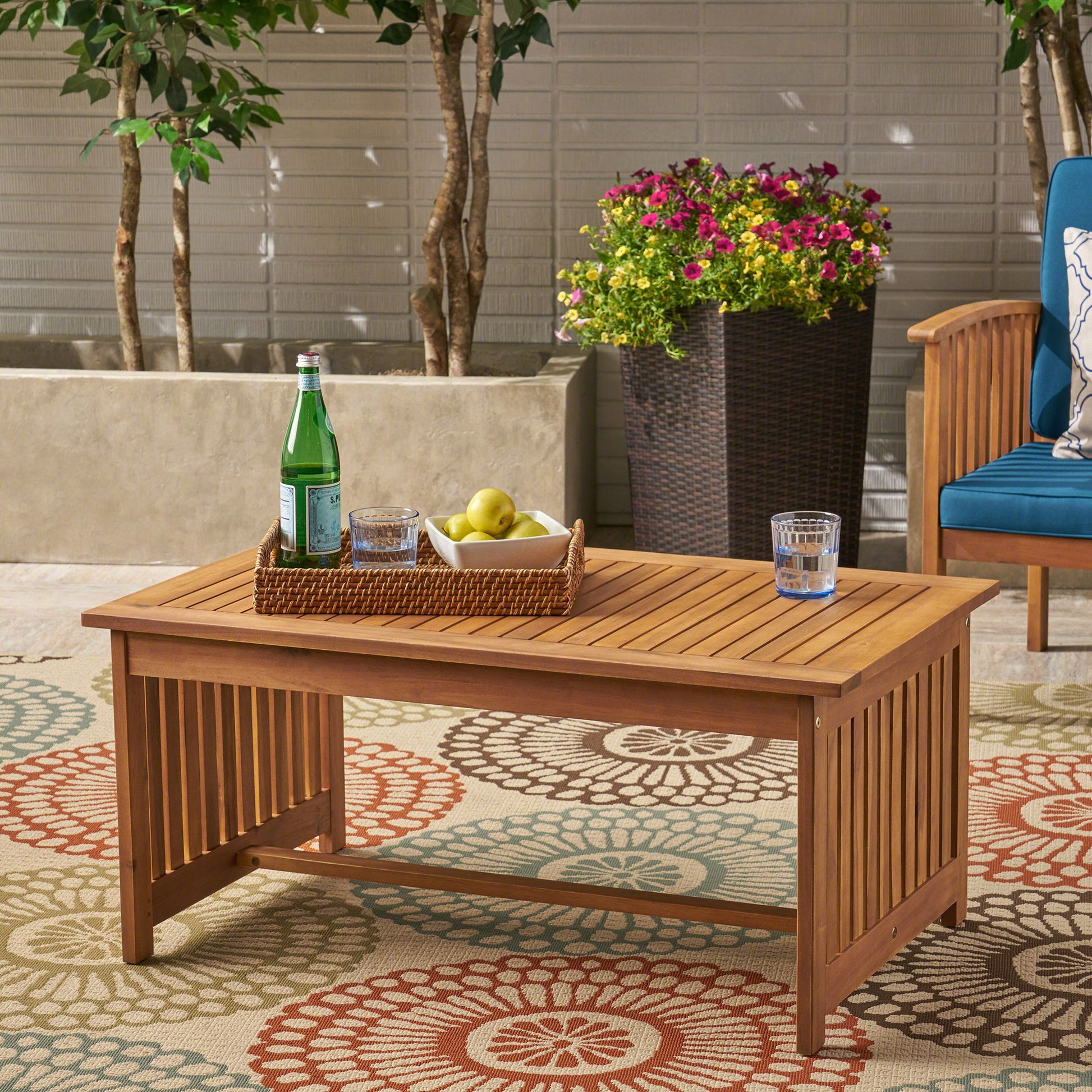 The Acacia Wood Rectangular Patio Coffee Table for Outside sits on a patterned rug, holding a basket with a water bottle, apples, and a glass of water. Potted plants and a blue cushioned chair appear in the background.