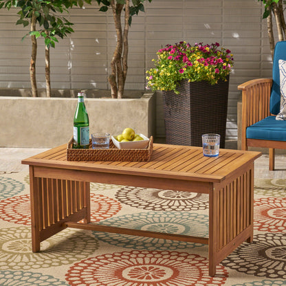 The Acacia Wood Rectangular Patio Coffee Table for Outside sits on a patterned rug, holding a basket with a water bottle, apples, and a glass of water. Potted plants and a blue cushioned chair appear in the background.
