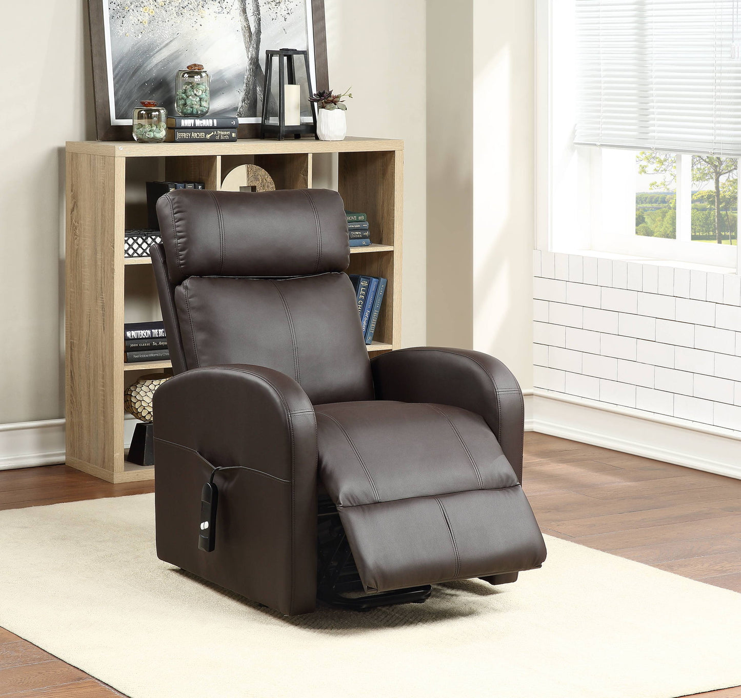 The Ricardo Power Motion Recliner With Lift, upholstered in dark brown leather, is placed on a light rug in a wood-floored living room with a bookshelf and a window letting in natural light.