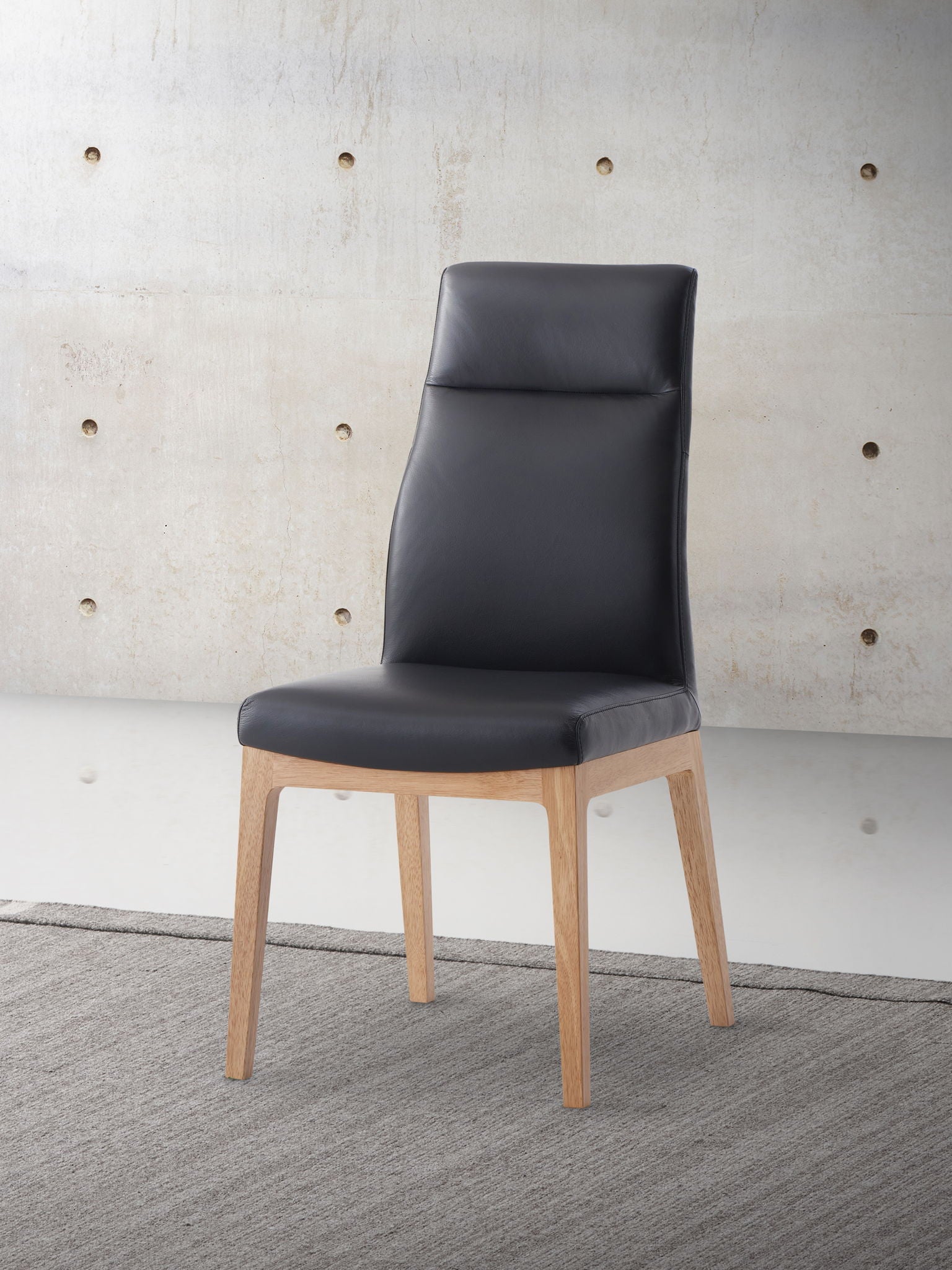 The Raquan - Leather Side Chair (Set of 2) - Black Pu features a black cushioned seat and back, light wood legs, and is shown on a gray rug before a beige concrete wall with circular indentations.