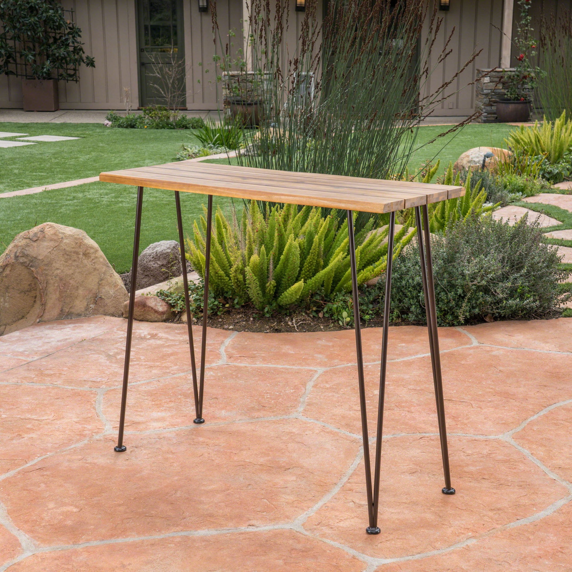 The Outdoor Bar Table With Wood Tabletop And Iron Hairpin Legs - Black / Teak sits on a stone patio in a landscaped garden with green plants, rocks, and a brown building in the background.