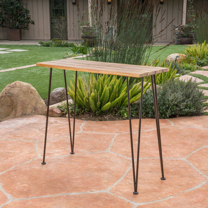 The Outdoor Bar Table With Wood Tabletop And Iron Hairpin Legs - Black / Teak sits on a stone patio in a landscaped garden with green plants, rocks, and a brown building in the background.