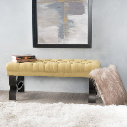 The Hilton Ottoman Bench Classic Style in yellow, featuring tufted upholstery and curved dark legs, sits beneath an abstract painting. Two books rest on the bench, while a beige pillow lies on a white rug nearby against light walls and flooring.