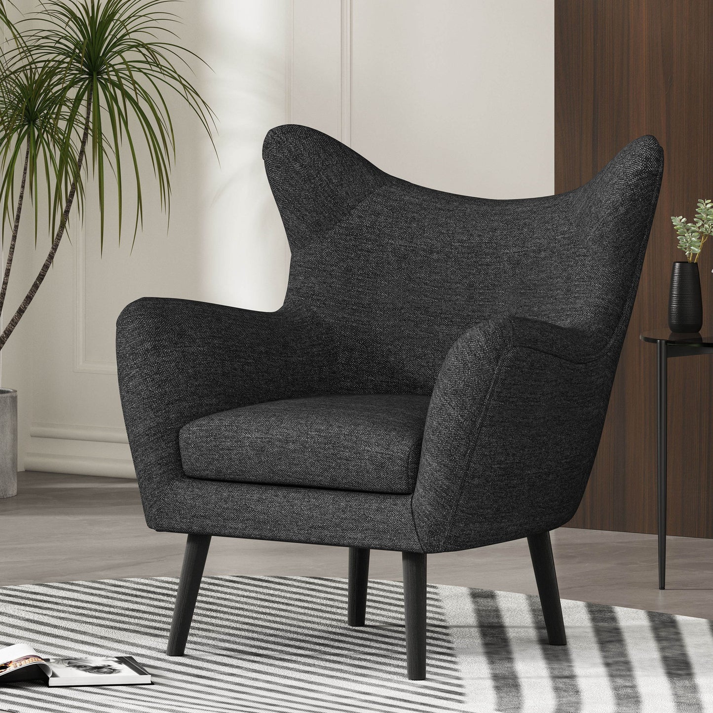 A Mid-Century Modern Arm Chair with wingback design and wood legs in dark gray sits on a striped rug in a stylish living room, next to a tall potted plant and a small black side table.