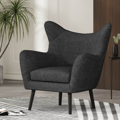 A Mid-Century Modern Arm Chair with wingback design and wood legs in dark gray sits on a striped rug in a stylish living room, next to a tall potted plant and a small black side table.