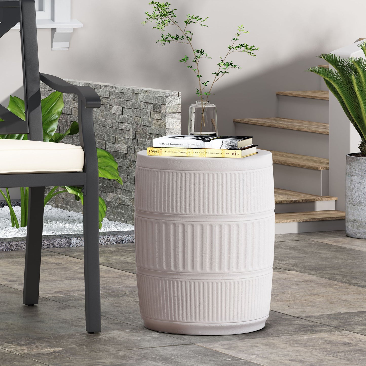 The Outdoor Patio End Table in Antique White, with a ribbed cylindrical design, holds books, a glass of water, and a vase with green branches beside a cushioned chair near indoor plants and modern stone steps.