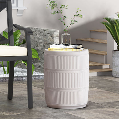 The Outdoor Patio End Table in Antique White, with a ribbed cylindrical design, holds books, a glass of water, and a vase with green branches beside a cushioned chair near indoor plants and modern stone steps.