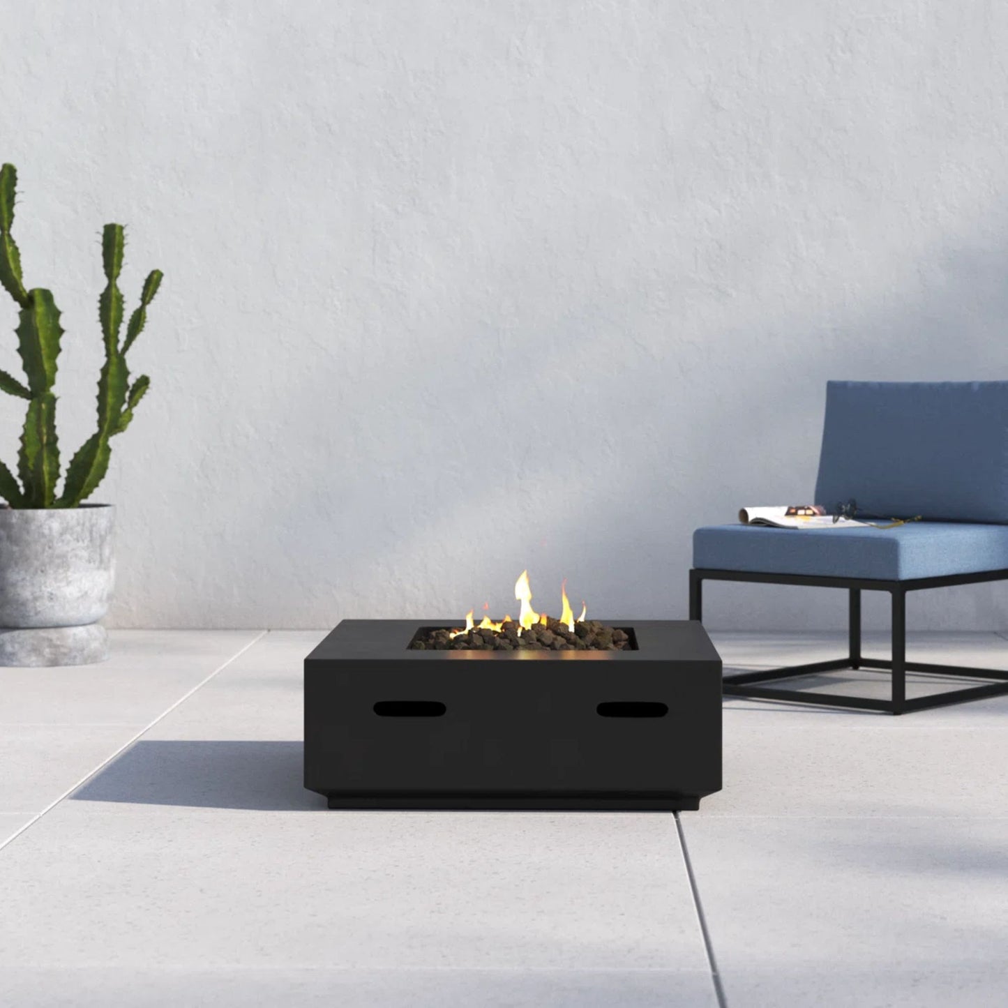 A modern patio features the Reinforced - Propane/Gas Fire Pit Table, a blue cushioned chair with a black metal frame, and a tall potted cactus set against a light gray wall and tiled floor.