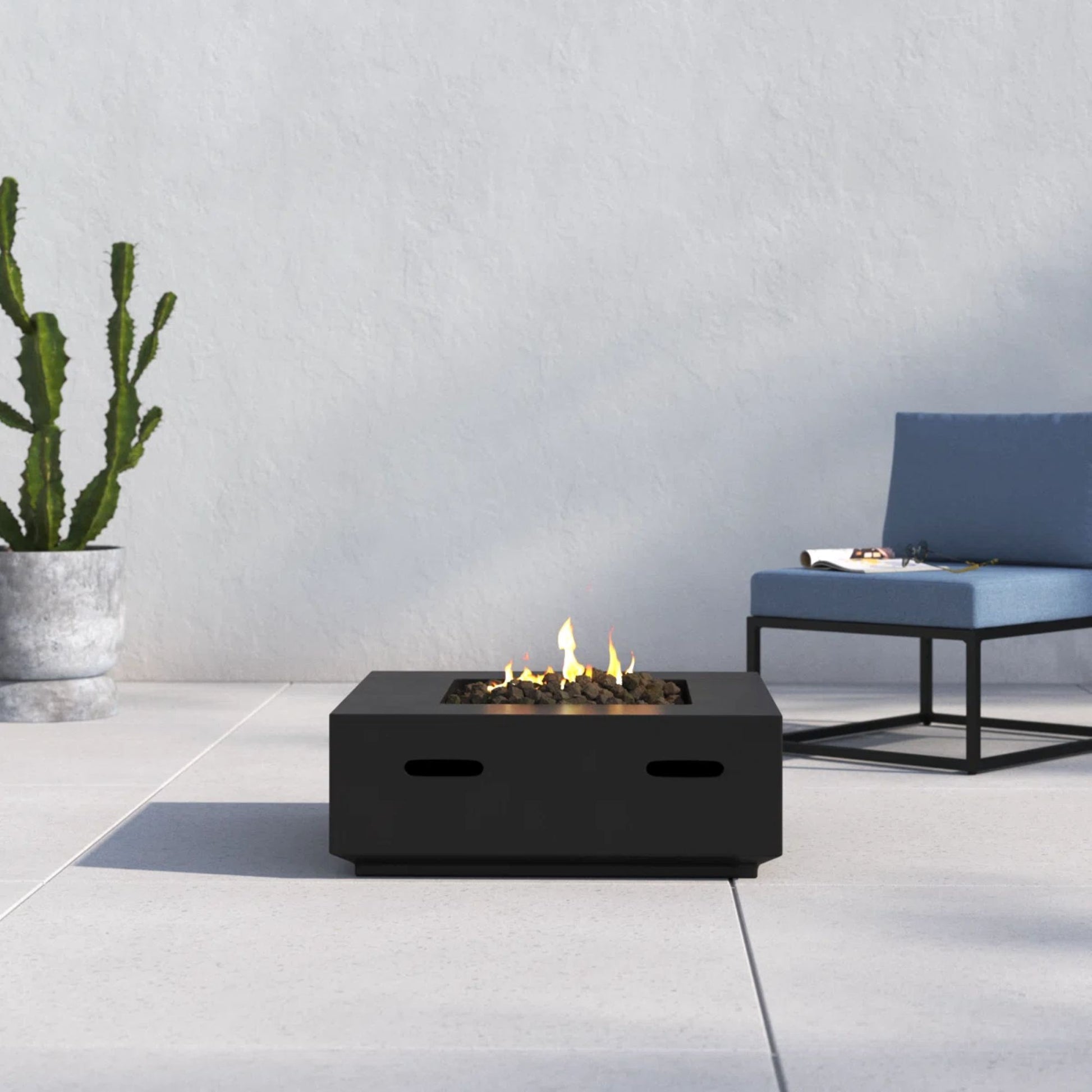 A modern patio features the Reinforced - Propane/Gas Fire Pit Table, a blue cushioned chair with a black metal frame, and a tall potted cactus set against a light gray wall and tiled floor.