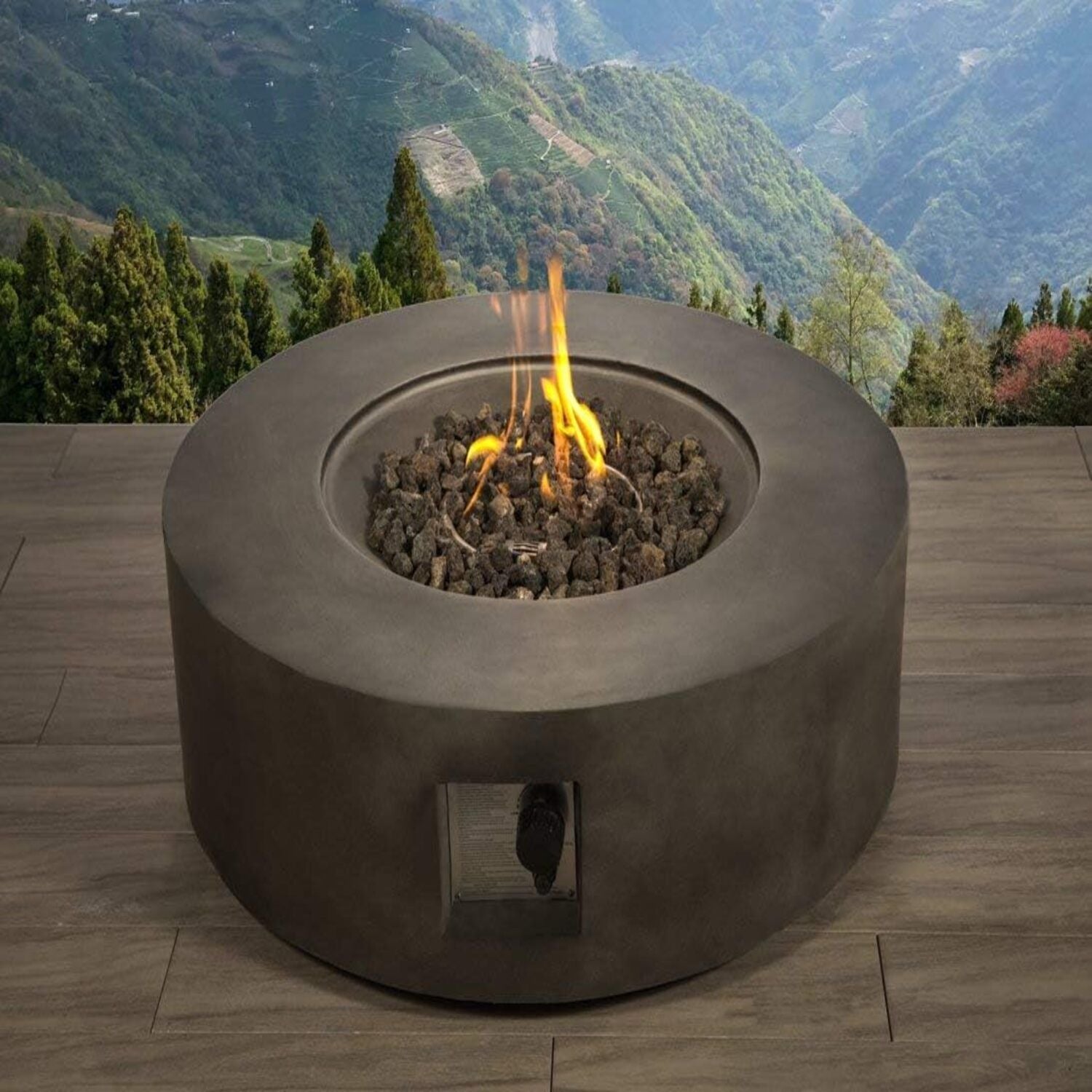 The Fibre Reinforced Outdoor Fire Pit Table - Charcoal, featuring flames and lava rocks, is placed on a wooden deck that overlooks a scenic mountain landscape filled with green trees and valleys.