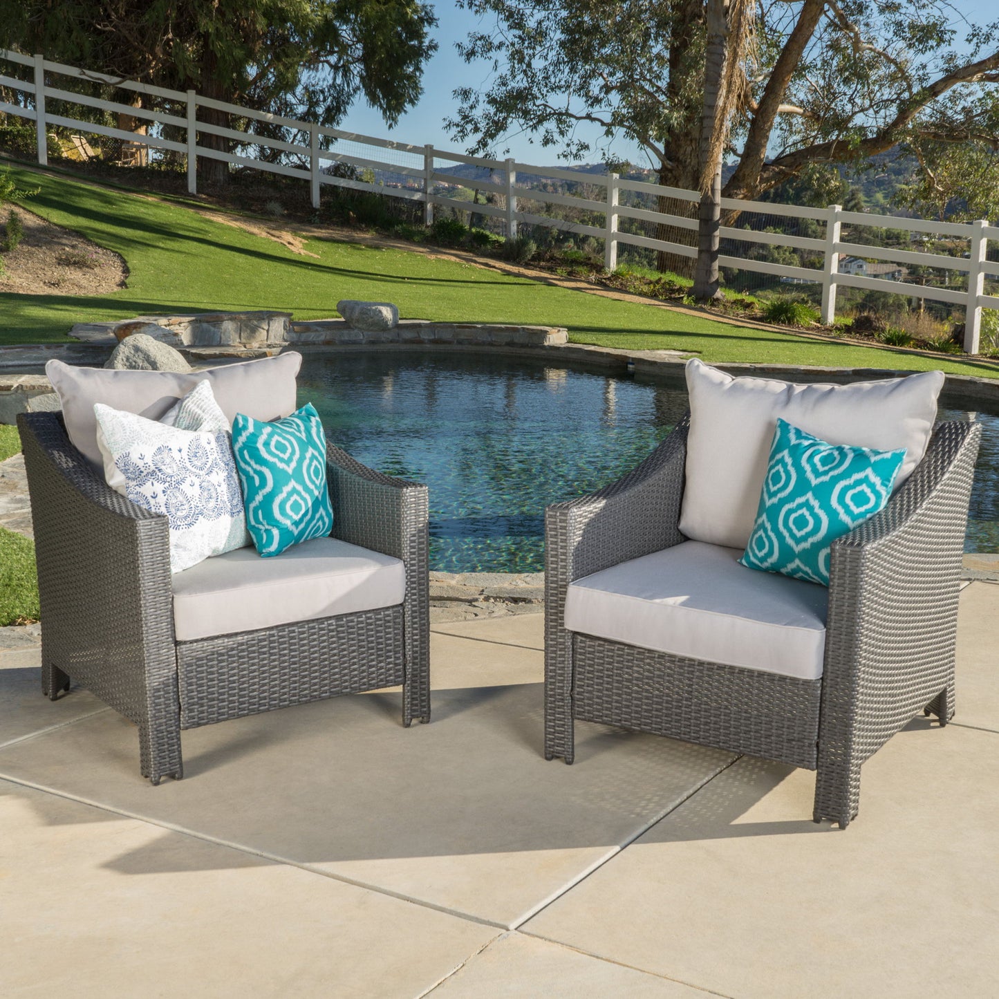 Two Antibes Armchairs with water-resistant cushions and blue patterned plus white pillows sit on a patio beside a small pool, surrounded by grass, rocks, trees, and a white fence.