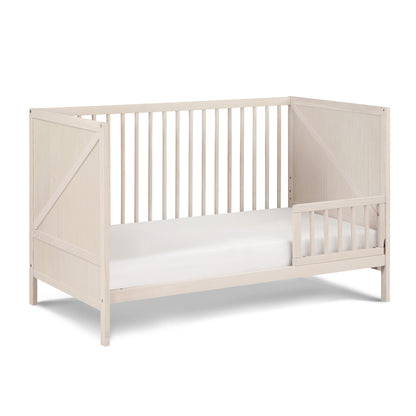 The Pixie Zen 3-in-1 Crib in Walnut features slatted sides, a minimalist light wood design, and includes one half-length right-side guardrail with a white mattress.