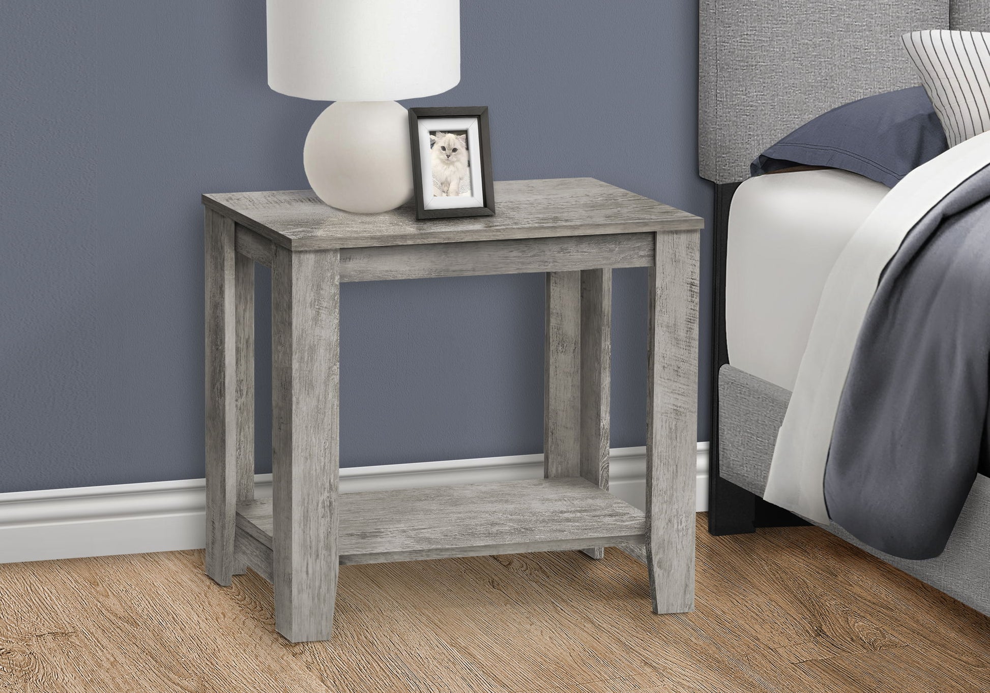 An Accent Table, Side Transitional with an elegant design stands beside a bed against a blue wall and wooden floor, topped with a white lamp and a framed photo.