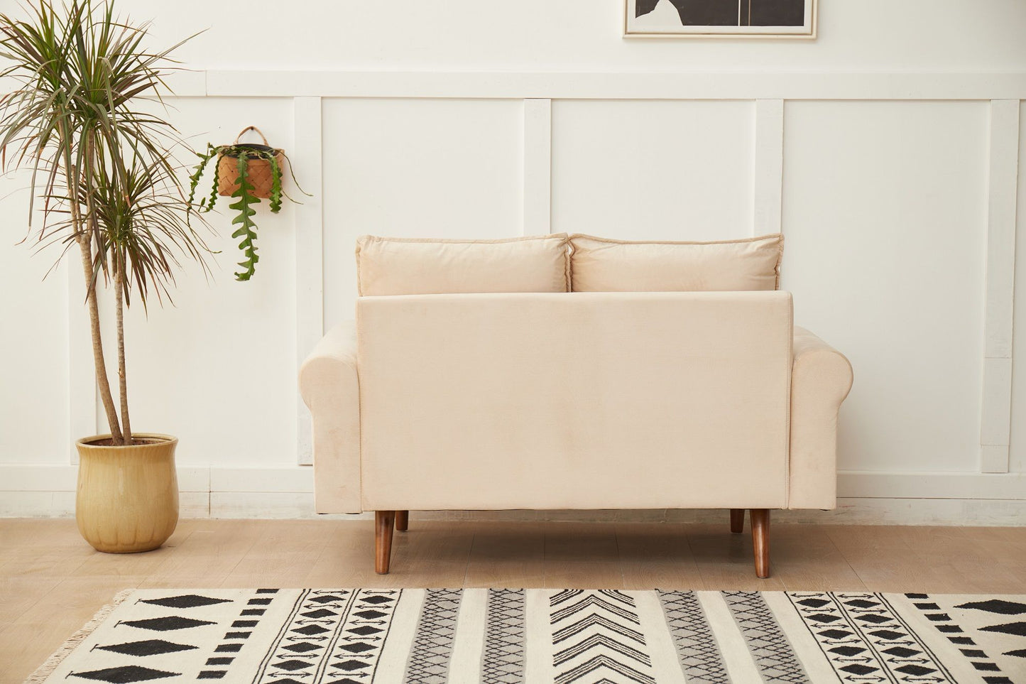 The Graceful Design Plush Comfort Loveseat sits against a white paneled wall, with a potted plant on the left, a hanging plant above, and a patterned black and white rug on the floor in front of it.