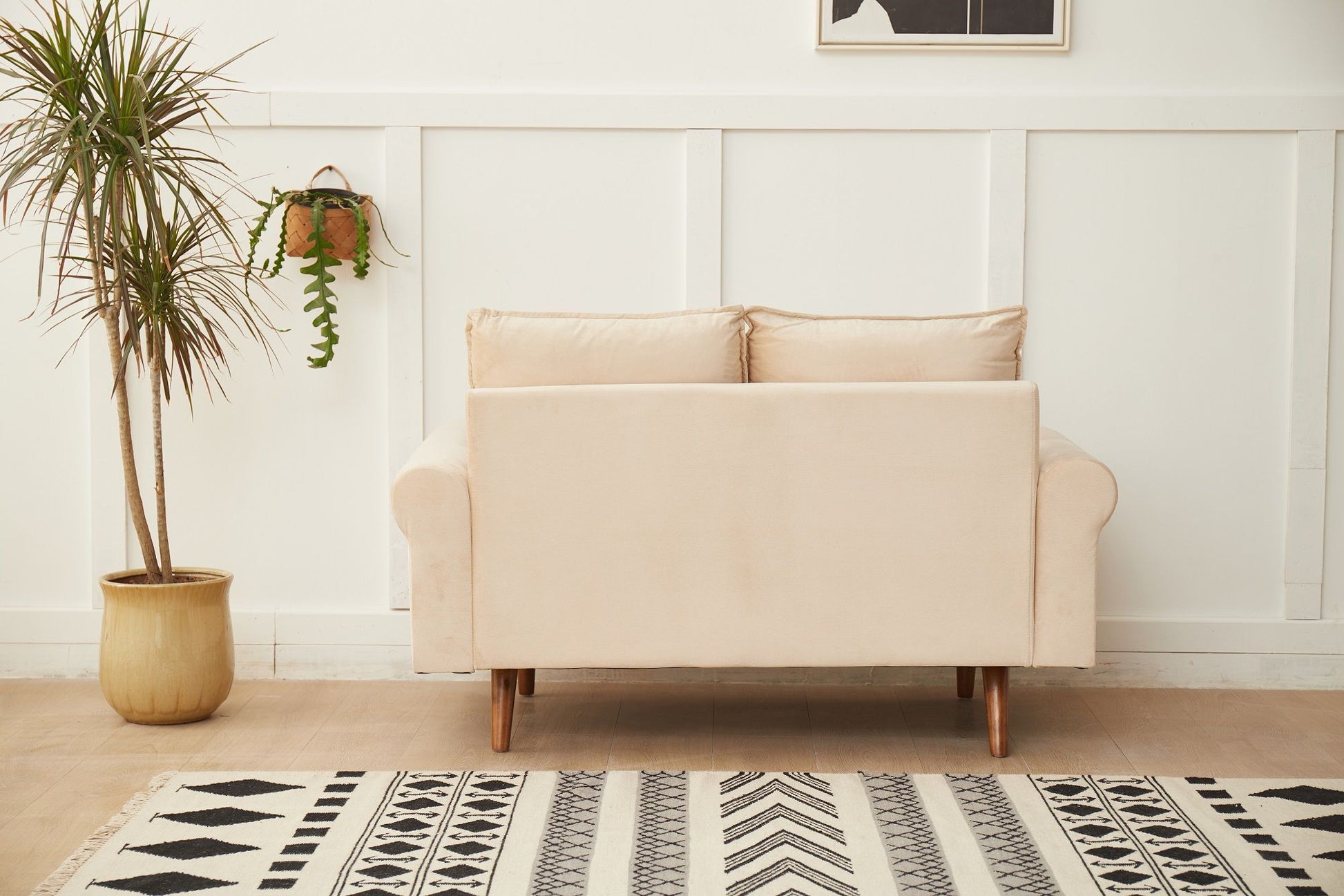 The Graceful Design Plush Comfort Loveseat sits against a white paneled wall, with a potted plant on the left, a hanging plant above, and a patterned black and white rug on the floor in front of it.