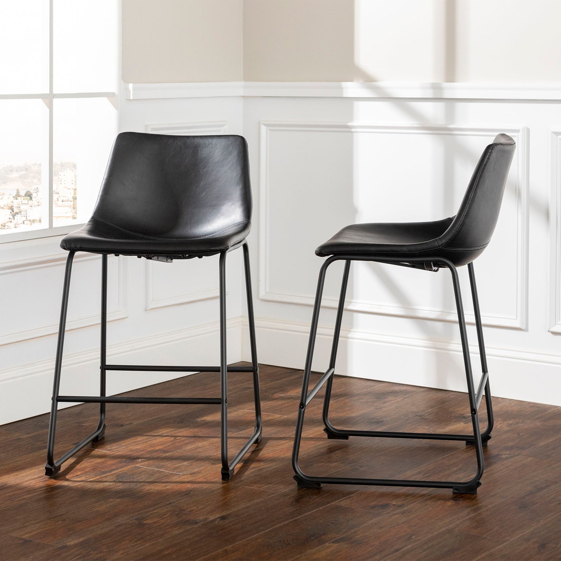 A set of two Industrial Counter Stools with metal legs in black sits on a dark wooden floor in a bright room featuring white paneled walls and large windows.