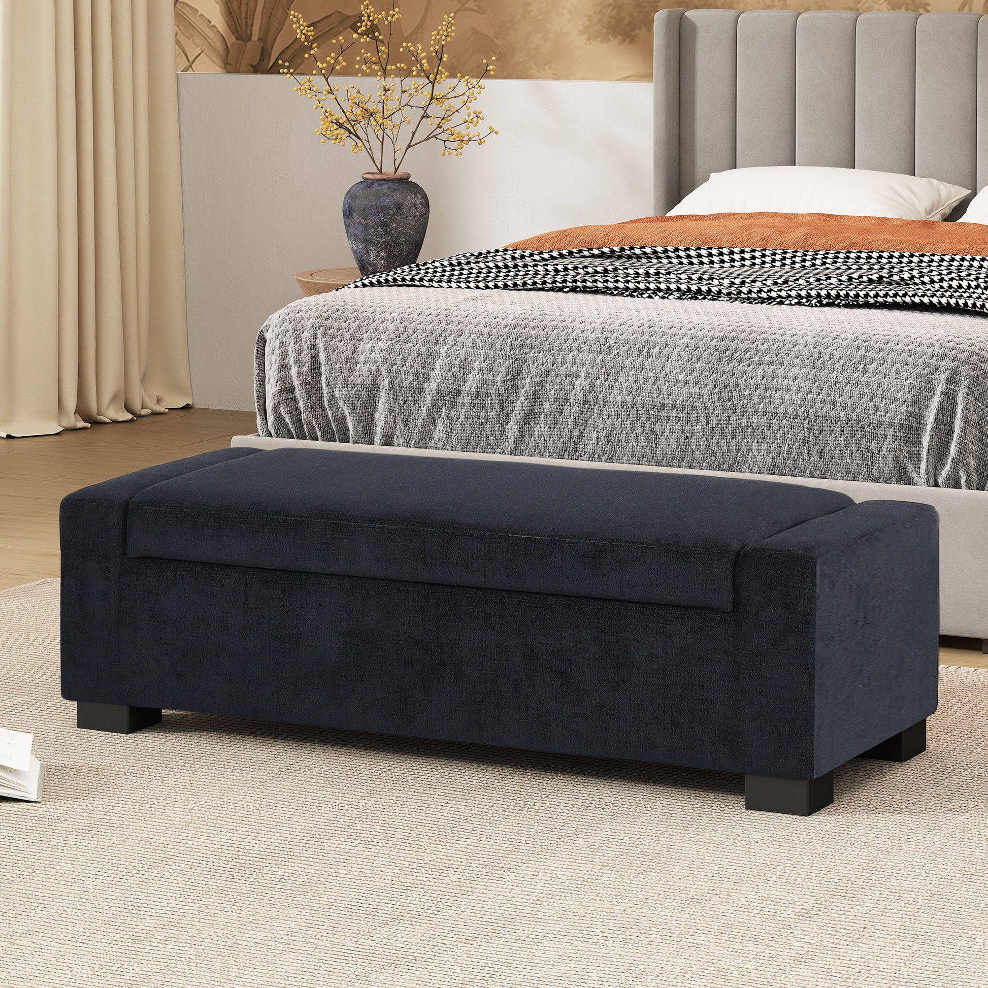 A Textured Fabric Ottoman Bench With Hinged Lid sits at the foot of a bed in a modern bedroom featuring neutral decor, a gray comforter, and a side table with a vase of yellow branches.