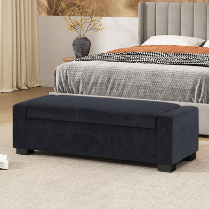 A Textured Fabric Ottoman Bench With Hinged Lid sits at the foot of a bed in a modern bedroom featuring neutral decor, a gray comforter, and a side table with a vase of yellow branches.