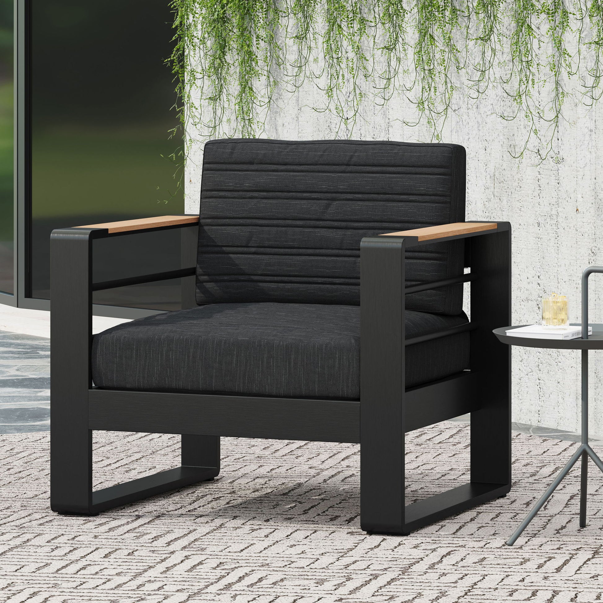 The Giovanna Club Chair Set features a modern black aluminum frame, dark cushioned seat and backrest, and wooden armrests. Placed on a patterned rug beside a small table, green vines cascade down a concrete wall in the background.