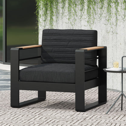 The Giovanna Club Chair Set features a modern black aluminum frame, dark cushioned seat and backrest, and wooden armrests. Placed on a patterned rug beside a small table, green vines cascade down a concrete wall in the background.