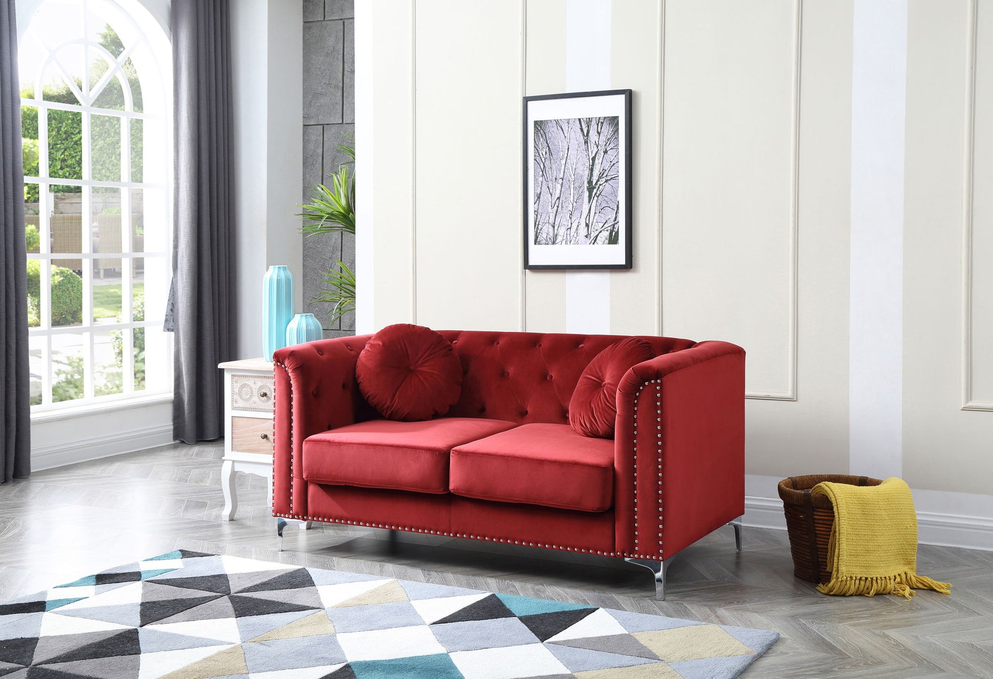 A modern living room featuring the Stylish Sloped Arm Loveseat in red velvet, two round cushions, a geometric rug, a white side table with vases, a basket with a yellow blanket, large windows, and a framed tree photo on the wall.