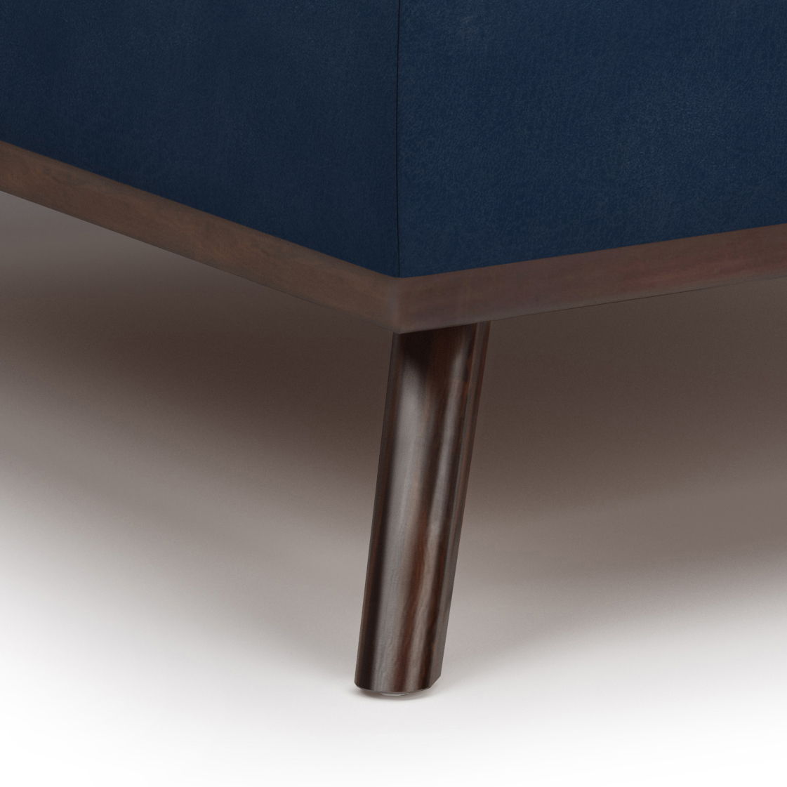 Close-up of a wooden leg supporting the corner of the Owen - Multifunctional Square Coffee Table Storage Ottoman with dark blue upholstery, wooden trim, on a light-colored floor.