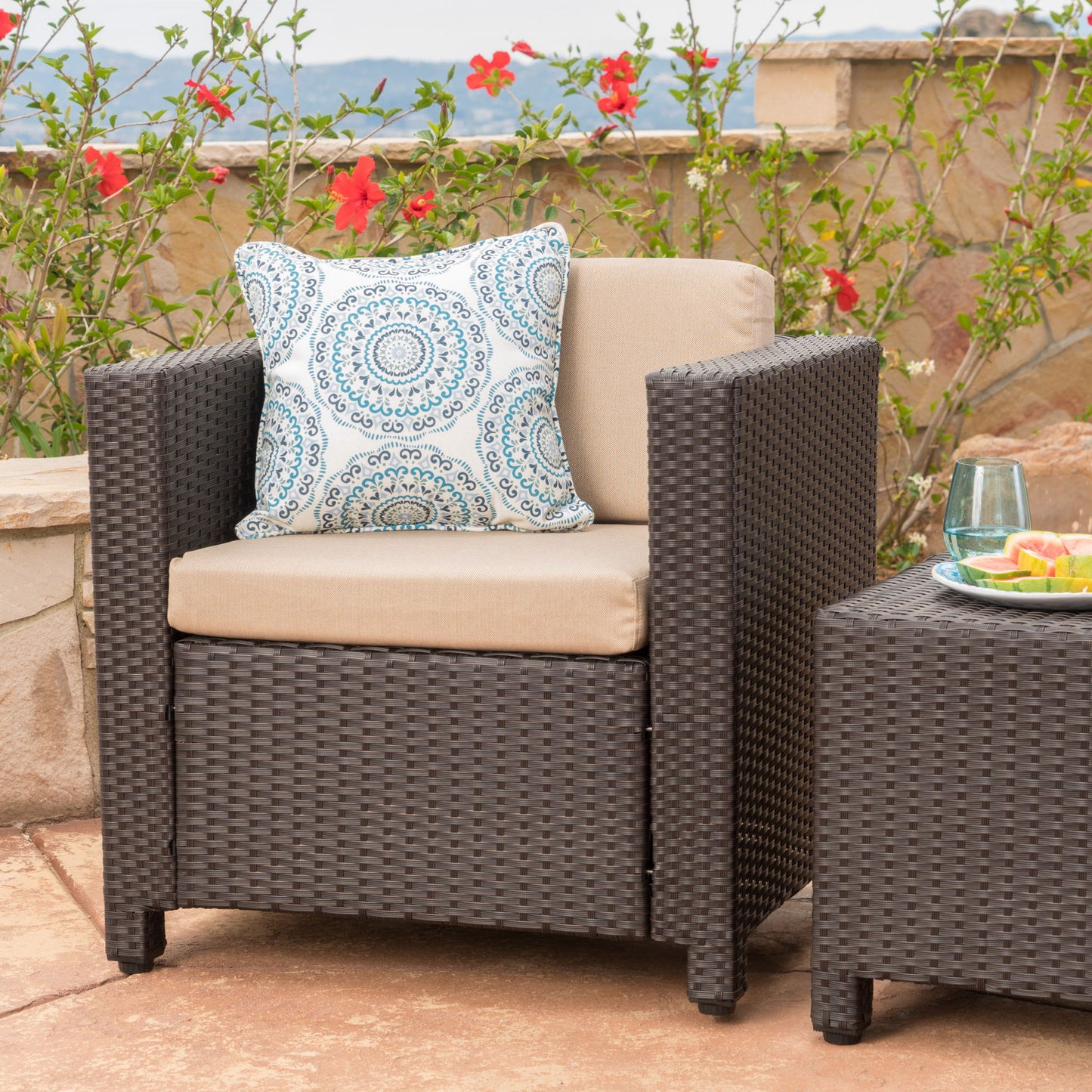 The Stylish Outdoor Club Chair With Weather Resistant Cushions is placed beside a matching table on a stone patio, accented with a blue patterned pillow, and set against blooming red flowers for an inviting outdoor scene.