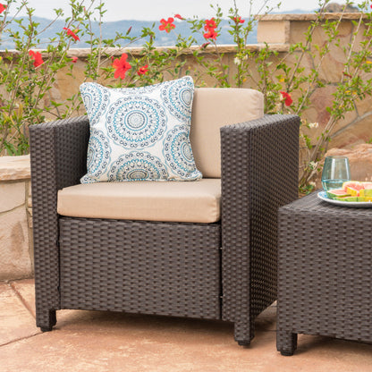 The Stylish Outdoor Club Chair With Weather Resistant Cushions is placed beside a matching table on a stone patio, accented with a blue patterned pillow, and set against blooming red flowers for an inviting outdoor scene.