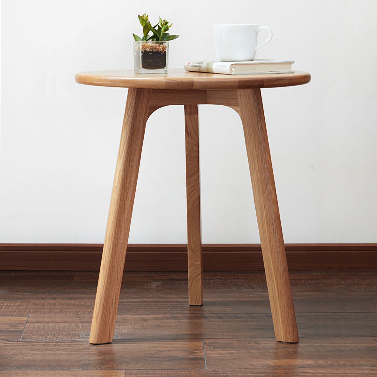 The Round Small End Table, Side Table For Bedroom & Balcony stands on three legs atop a wooden floor, holding a potted plant, two closed books, and a white cup with saucer. A plain white wall forms the background.