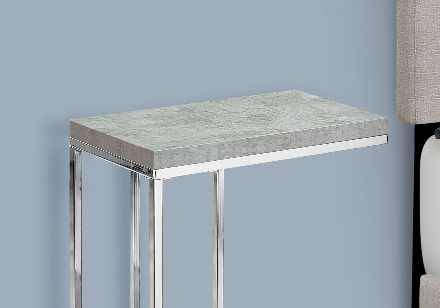 A contemporary accent table with a stylish C-shaped design and tempered glass top, featuring sleek metal legs, is placed by a beige upholstered surface against a light blue wall.