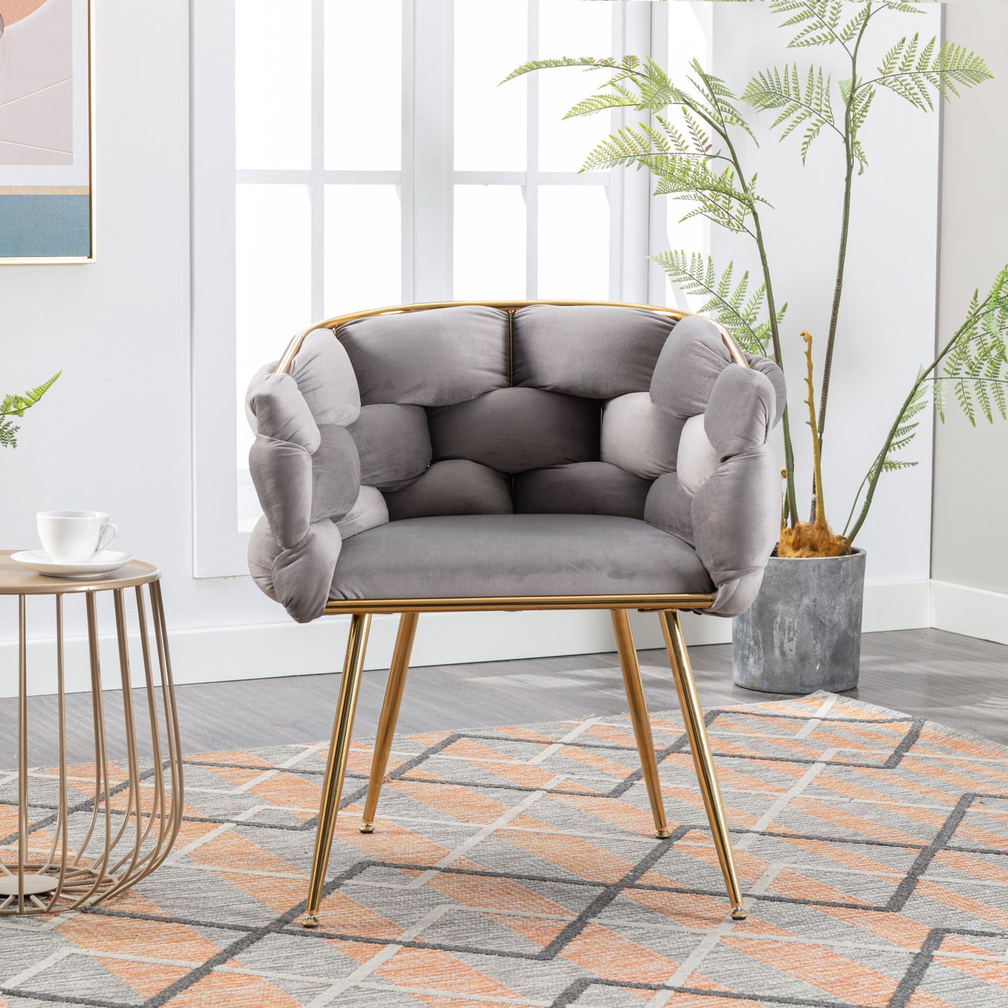 The Luxury Modern Simple Leisure Velvet Single Sofa Chair with gold legs sits on a patterned rug by a round side table with a cup and saucer. Tall plants and large windows in the background create a bright, stylish space.