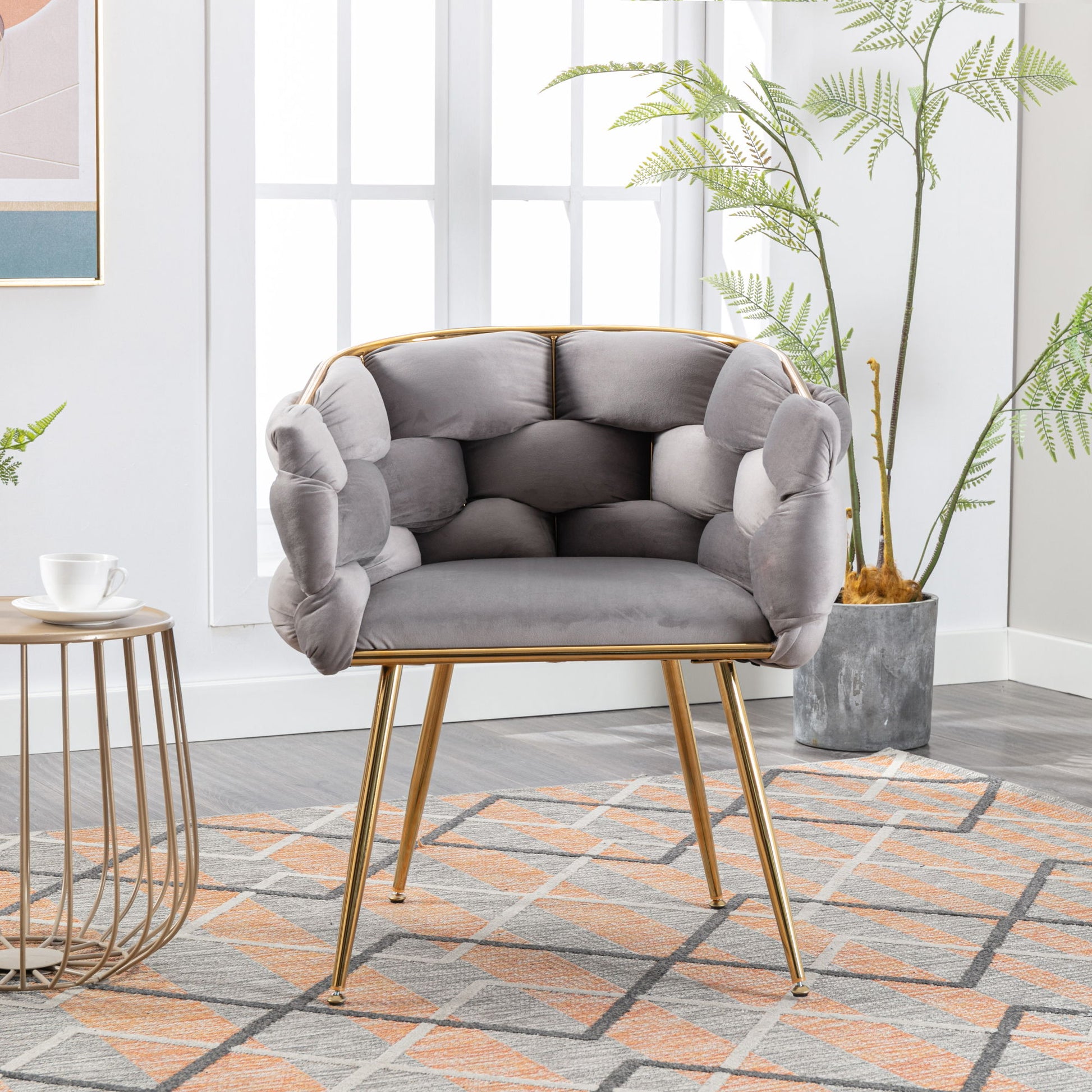 The Luxury Modern Simple Leisure Velvet Single Sofa Chair with gold legs sits on a patterned rug by a round side table with a cup and saucer. Tall plants and large windows in the background create a bright, stylish space.