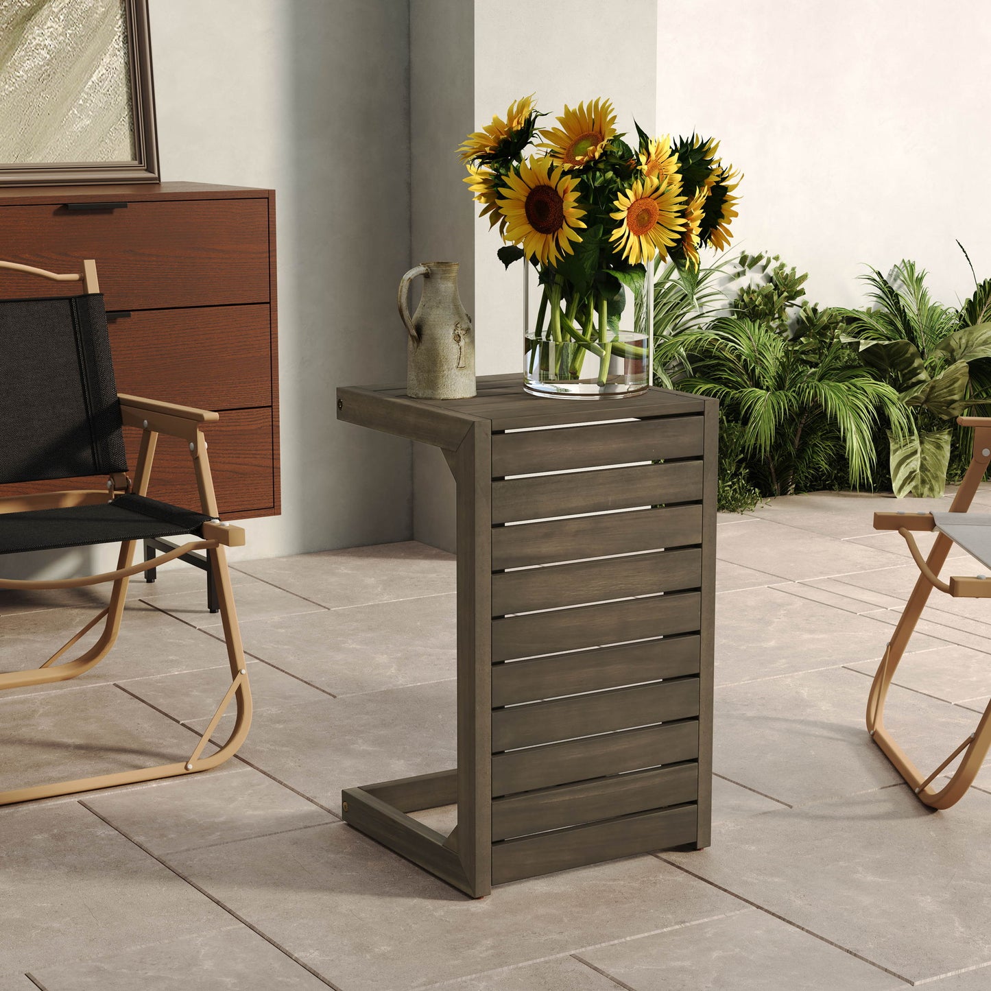 An Aluminum Outdoor C-Shaped Side Table for patio holds a glass vase of sunflowers and a ceramic jug, set on a tile-floored patio with a folding chair, cabinet, and lush green plants surrounding the area.