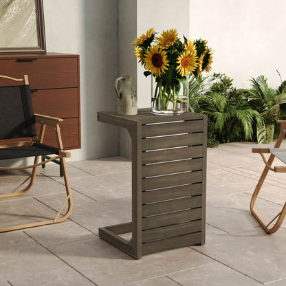 An Aluminum Outdoor C-Shaped Side Table for patio holds a glass vase of sunflowers and a ceramic jug, set on a tile-floored patio with a folding chair, cabinet, and lush green plants surrounding the area.