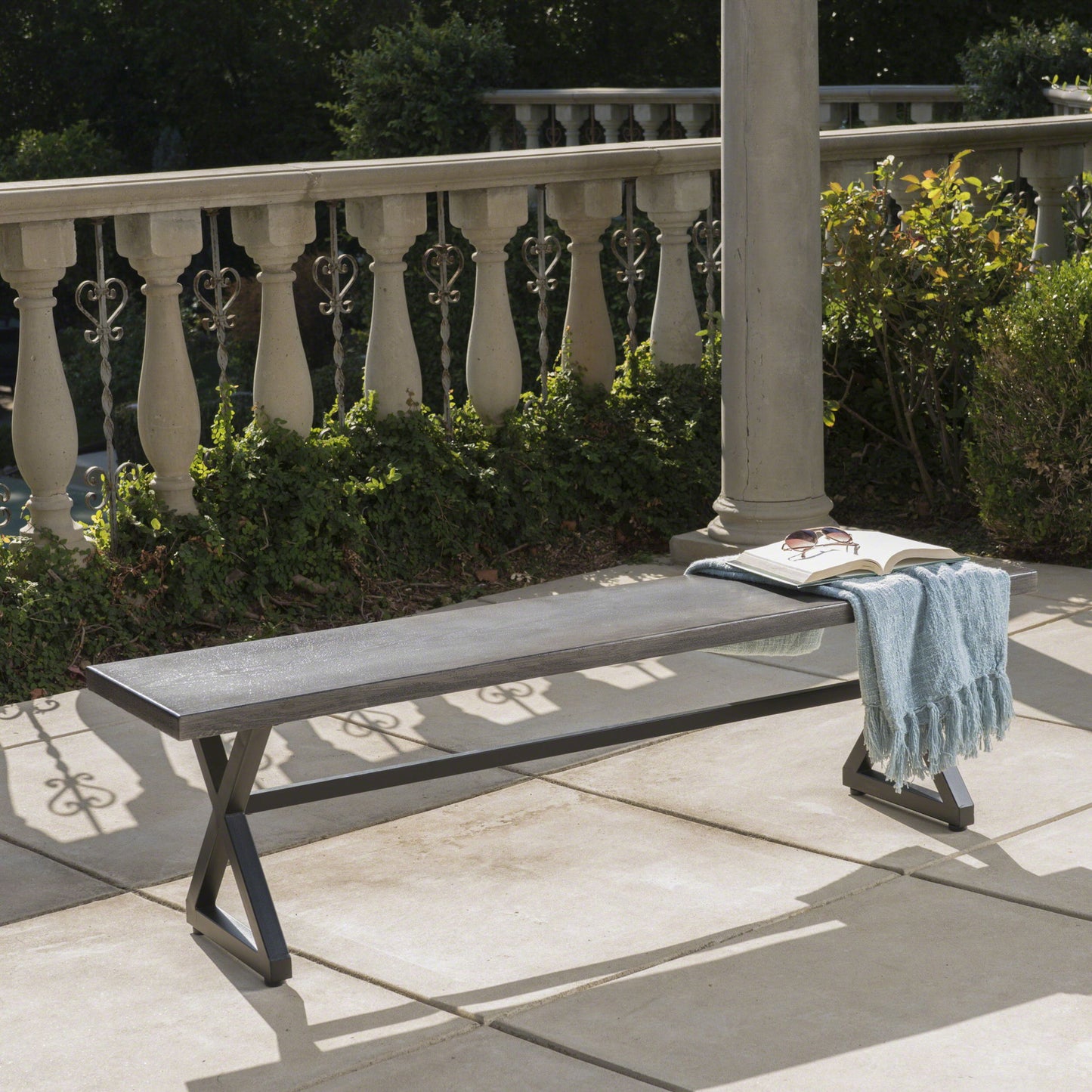 The Modern Aluminum Outdoor Dining Bench With X Base, featuring a metal frame and dark wood seat, sits on the patio with an open book, eyeglasses, and light blue scarf on top. Green shrubs and decorative railing complete the scene.