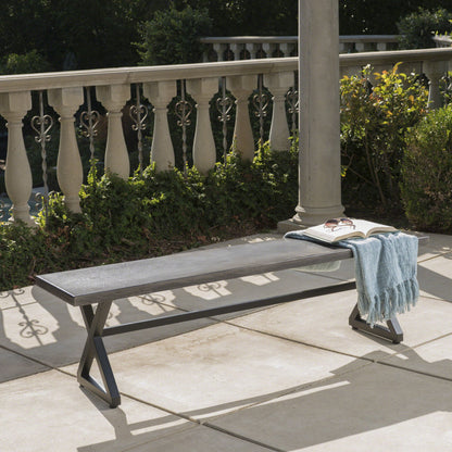 The Modern Aluminum Outdoor Dining Bench With X Base, featuring a metal frame and dark wood seat, sits on the patio with an open book, eyeglasses, and light blue scarf on top. Green shrubs and decorative railing complete the scene.