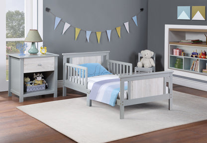 A modern toddler bedroom featuring the Connelly - Reversible Panel Toddler Bed in gray, blue and white bedding, a nightstand with a lamp and toys, a white teddy bear on a chair, shelves with books and toys, and pastel bunting on a gray wall.