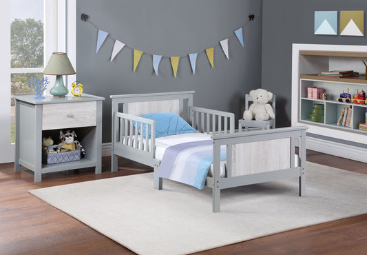 A modern toddler bedroom featuring the Connelly - Reversible Panel Toddler Bed in gray, blue and white bedding, a nightstand with a lamp and toys, a white teddy bear on a chair, shelves with books and toys, and pastel bunting on a gray wall.