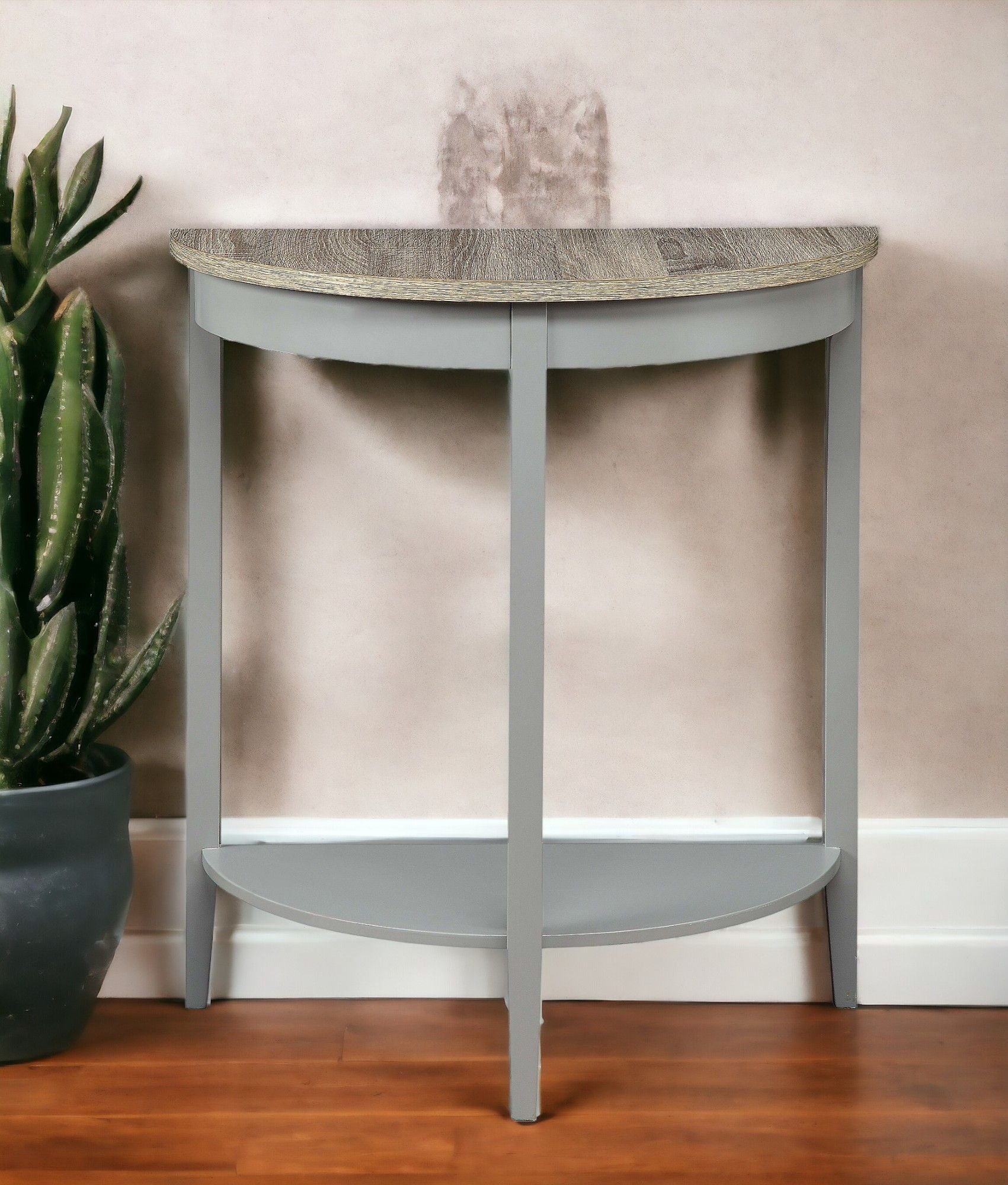 The Justino Console Table in gray and oak stands against a beige wall, with a green potted cactus next to it on the wooden floor. A faint stain is visible on the wall above the table.