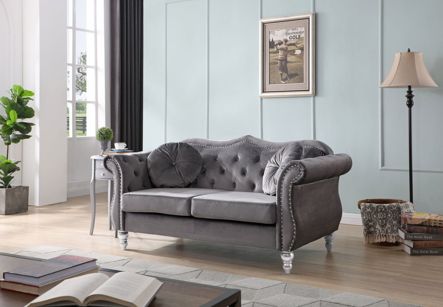 A modern living room features the Stylish Flared Arm Loveseat in gray velvet with tufted details, round pillows, a side table holding a plant, floor lamp, stacked books, and framed art on a pale blue wall. Sunlight streams through tall windows.