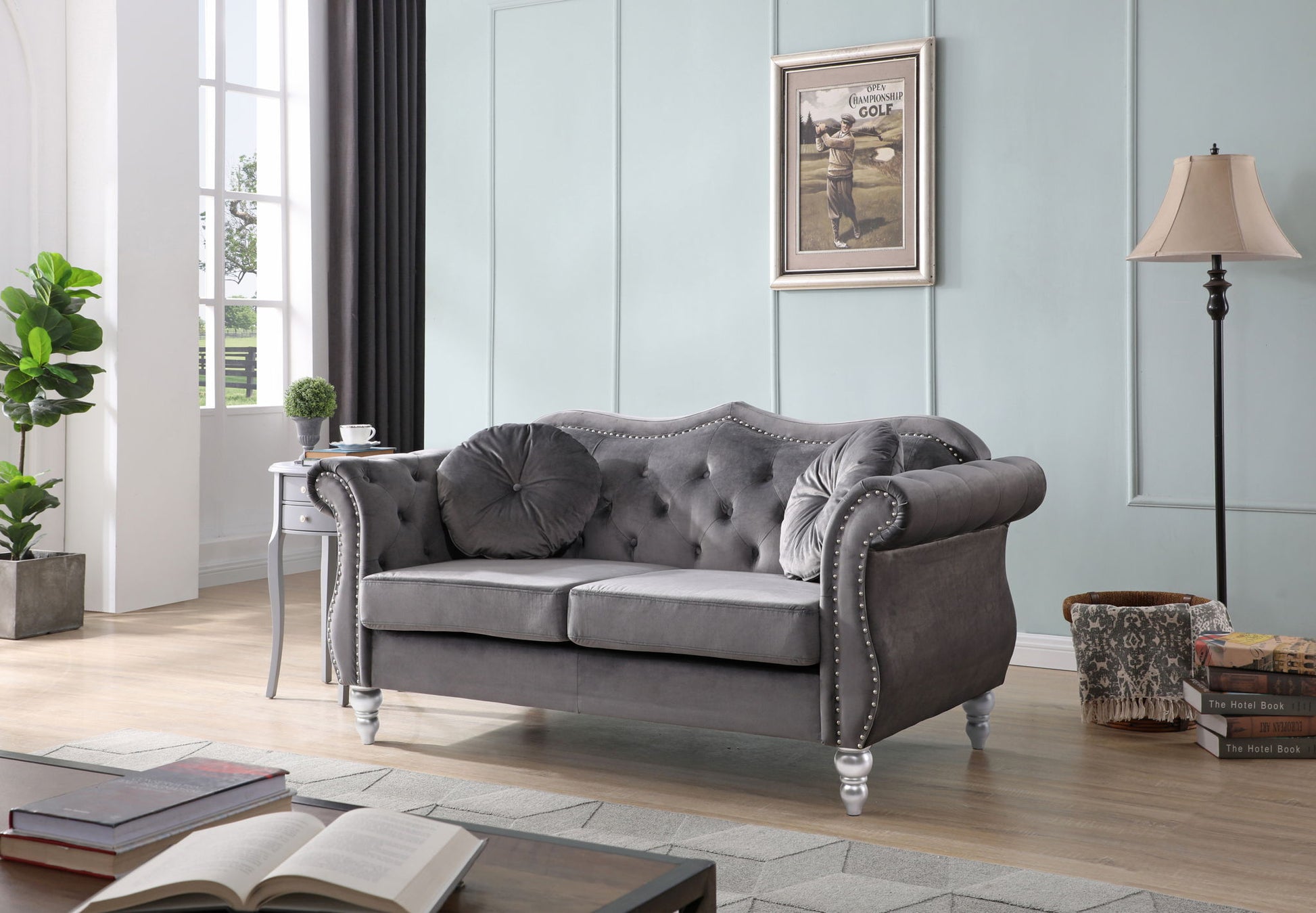 A modern living room features the Stylish Flared Arm Loveseat in gray velvet with tufted details, round pillows, a side table holding a plant, floor lamp, stacked books, and framed art on a pale blue wall. Sunlight streams through tall windows.