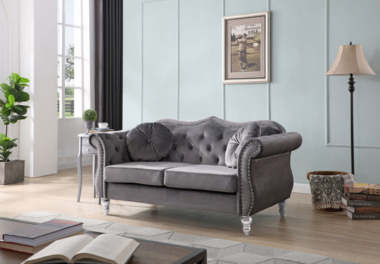A modern living room features the Stylish Flared Arm Loveseat in gray velvet with tufted details, round pillows, a side table holding a plant, floor lamp, stacked books, and framed art on a pale blue wall. Sunlight streams through tall windows.