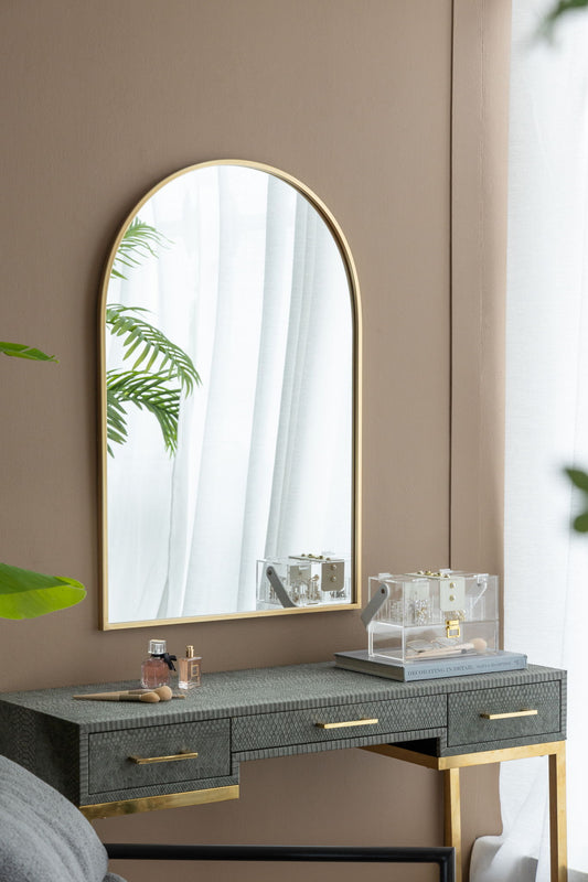 An Arched Mirror With Metal Frame hangs above a vanity with gold accents, holding perfume bottles and a clear jewelry box. The mirror reflects white curtains and greenery in the softly lit room.