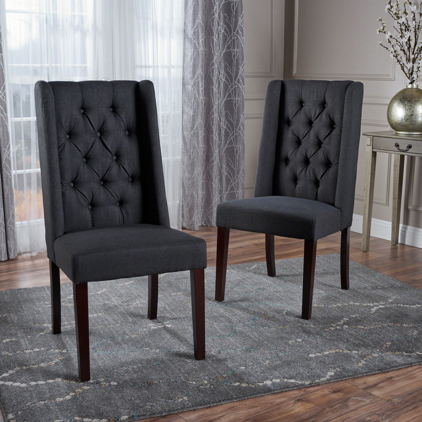 Two Sophisticated Tufted Dining Chairs With High Back Design and wooden legs rest on a gray patterned rug in a modern room featuring hardwood floors, sheer curtains, and stylish decorative accents.