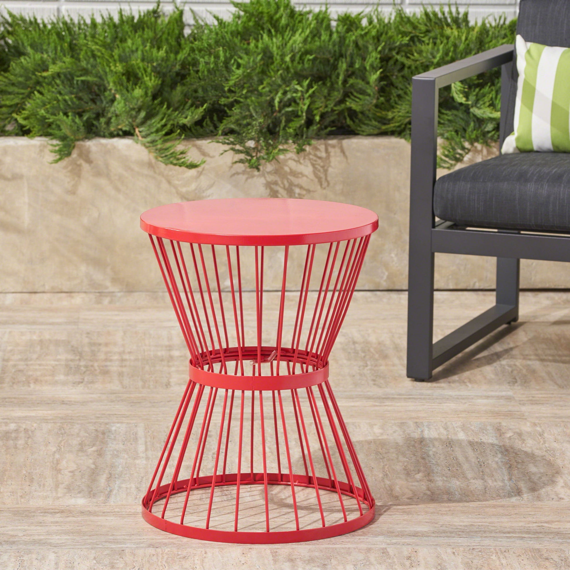 A modern outdoor space featuring the Lassen red metal hourglass side table beside a gray chair with a green and white striped pillow, set on a stone patio with lush green plants in the background.