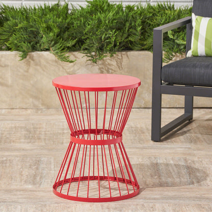 A modern outdoor space featuring the Lassen red metal hourglass side table beside a gray chair with a green and white striped pillow, set on a stone patio with lush green plants in the background.