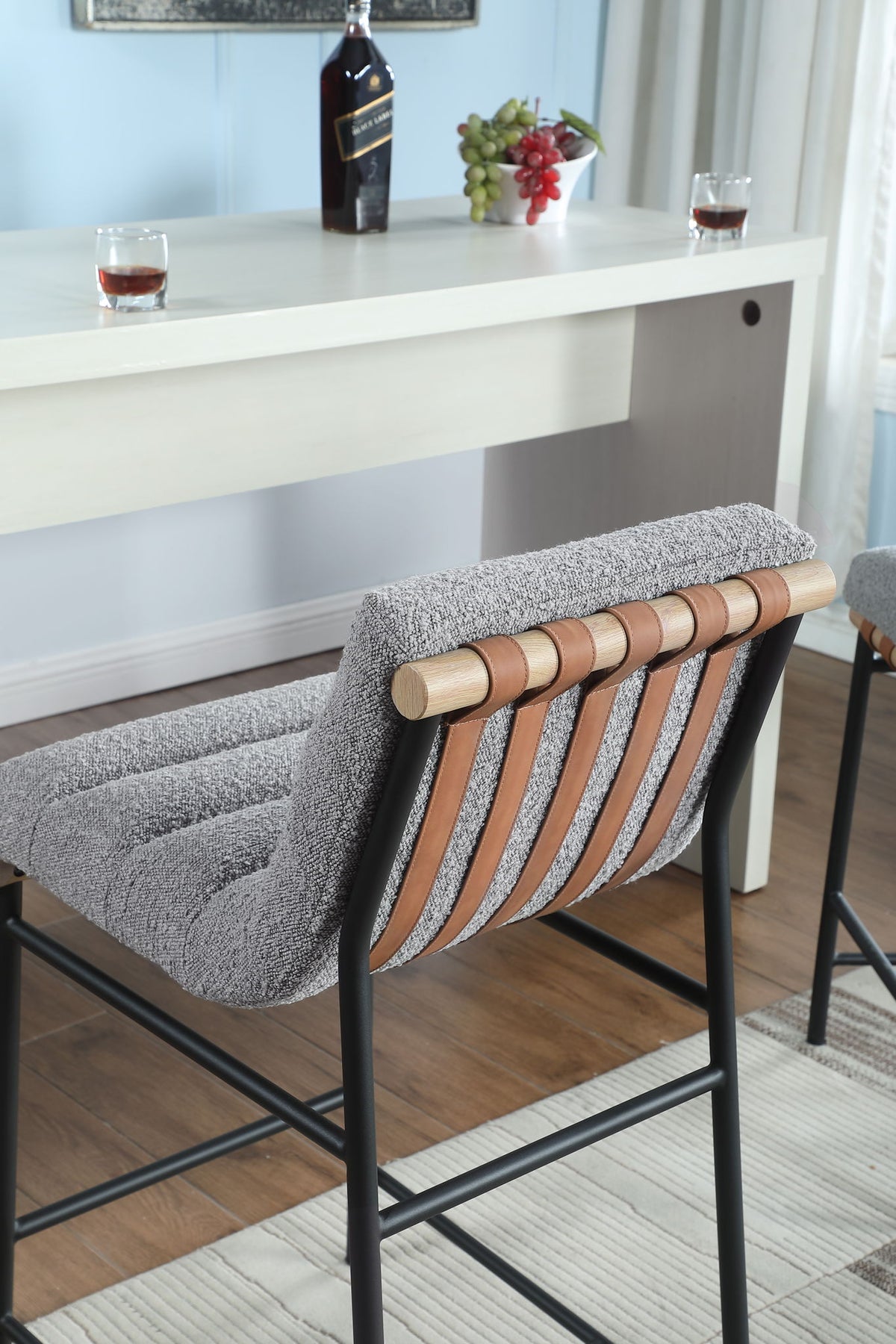 The Lahni - 17" Boucle Fabric Counter Height Chair with wooden and leather accents faces a white table topped with drinks, a bottle, and grapes. Sunlight streams in through a window with white curtains in the background.