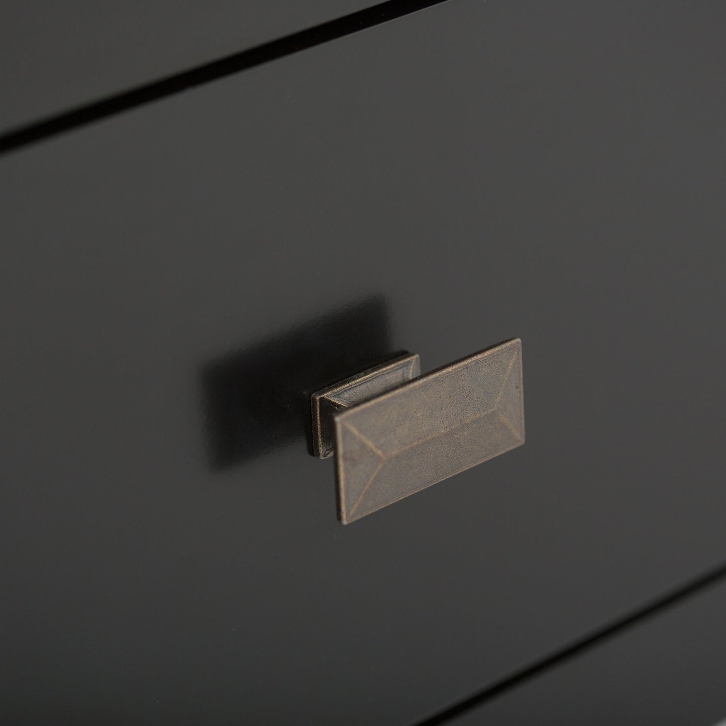 Close-up of a matte black drawer on the Artisan - Handcrafted, Tall TV Media Stand, featuring a rectangular bronze-colored handle with beveled edges and geometric detailing. The smooth surface subtly reflects the handle's shadow.