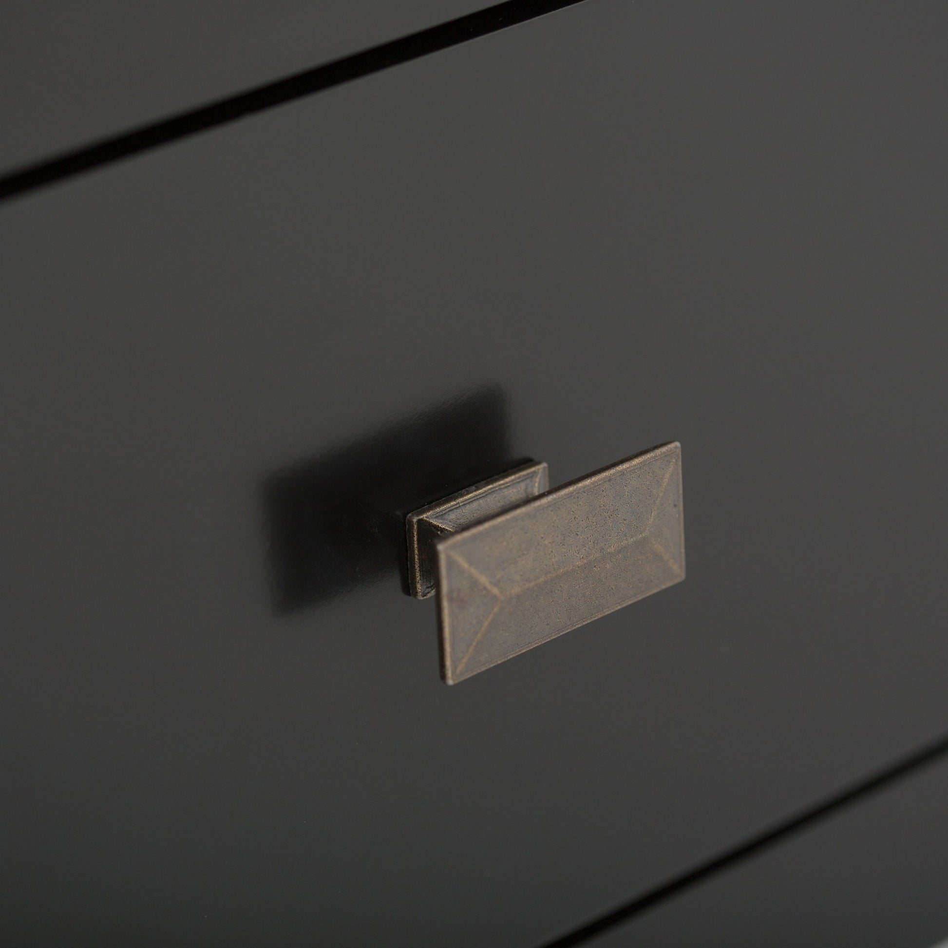 Close-up of a matte black drawer on the Artisan - Handcrafted, Tall TV Media Stand, featuring a rectangular bronze-colored handle with beveled edges and geometric detailing. The smooth surface subtly reflects the handle's shadow.
